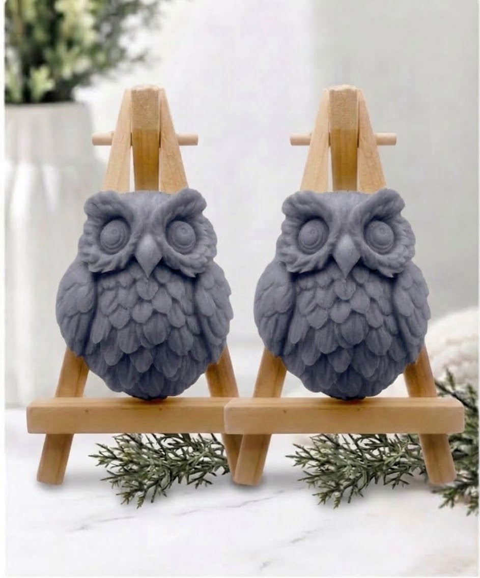Two owl-shaped handmade soaps in a charcoal colour displayed side by side on small wooden easel stands on a white marble surface.