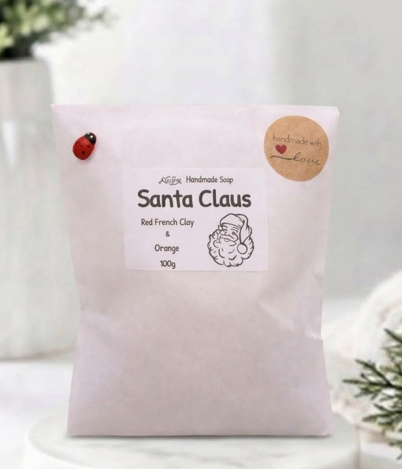 A 100g packet of Santa Claus handmade soap, made from red French clay and orange. The packet is light pink with a white label, a small ladybug decoration, and a round wooden badge, displayed on a white marble stand with soft-focus holiday decorations in the background.