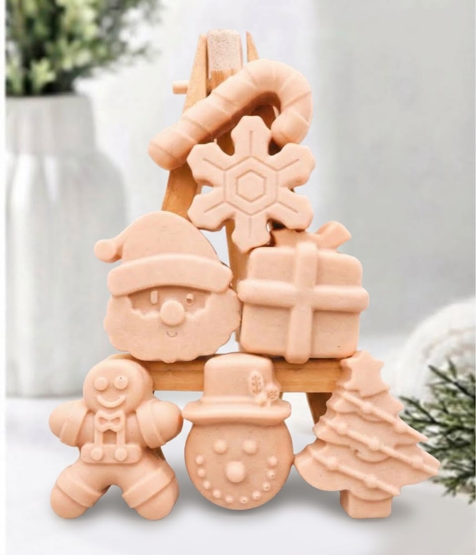 A festive collection of soft pink mini Christmas soaps, shaped as Santa, a snowman, a Christmas tree, a gingerbread man, a present, a snowflake, and a candy cane, arranged on a mini wooden easel.