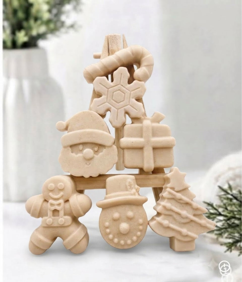 A festive collection of yellow coloured mini Christmas soaps, shaped as Santa, a snowman, a Christmas tree, a gingerbread man, a present, a snowflake, and a candy cane, arranged on a mini wooden easel.