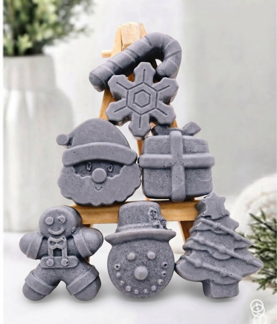 A festive collection of charcoal coloured Christmas soaps, shaped as Santa, a snowman, a Christmas tree, a gingerbread man, a present, a snowflake, and a candy cane, arranged on a mini wooden easel.