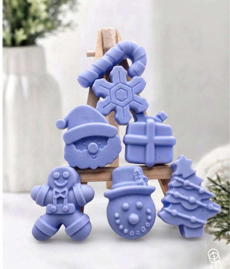 A set of lavender blue Christmas-themed soaps like a candy cane, snowflake, Santa face, gift box, gingerbread man, snowman, and Christmas tree, displayed on a small wooden easel against a soft, blurred indoor background.