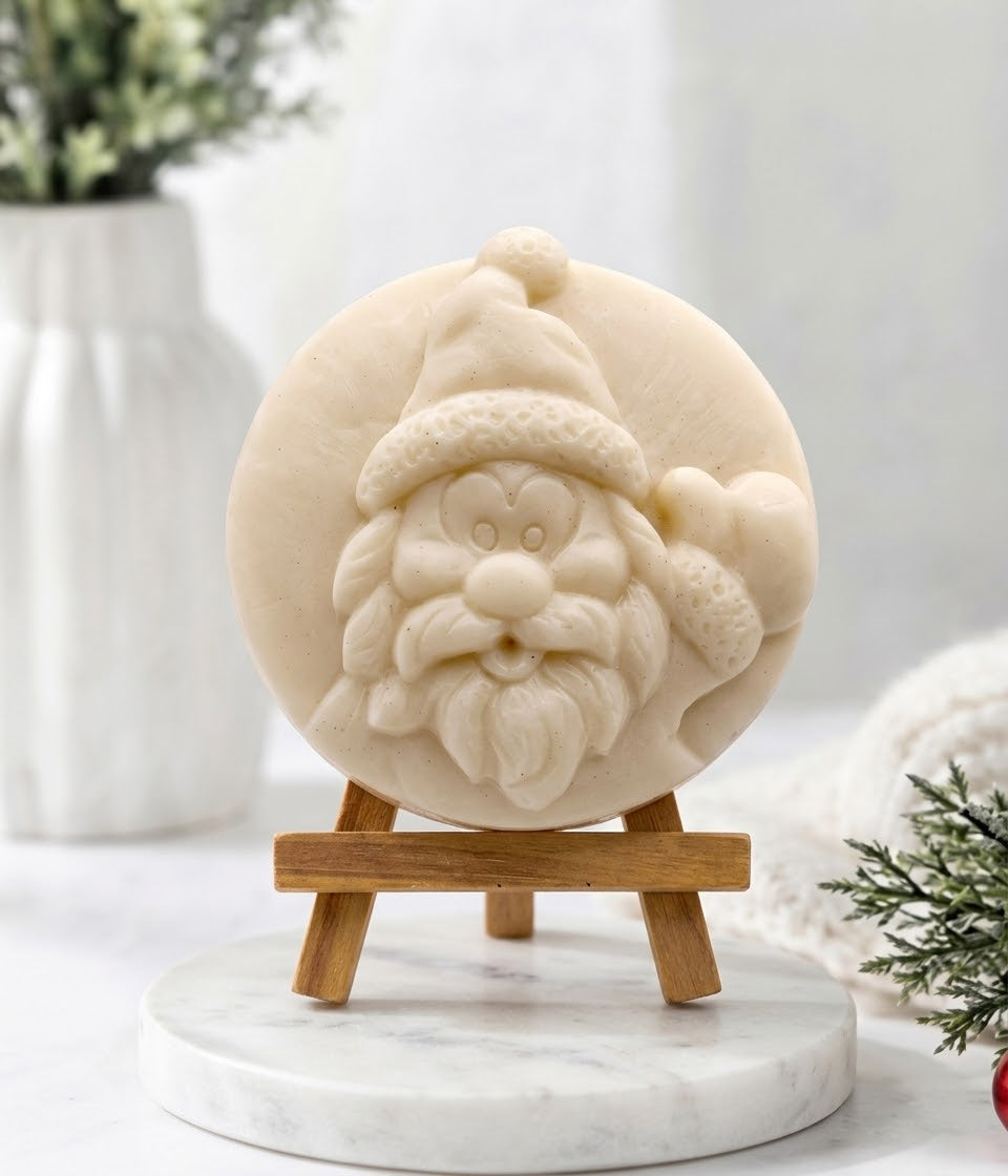 A close-up photograph of a circular, hand-carved soap disc featuring a relief of Santa Claus’s face, rendered in a pale cream-yellow colour. The soap rests on a small, light-wood tripod easel, which is positioned on a round white marble pedestal. The background is a soft-focus white setting with a vase and greenery, creating a clean, cosy display.