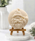 A close-up photograph of a circular, hand-carved soap disc featuring a relief of Santa Claus’s face, rendered in a pale cream-yellow colour. The soap rests on a small, light-wood tripod easel, which is positioned on a round white marble pedestal. The background is a soft-focus white setting with a vase and greenery, creating a clean, cosy display.
