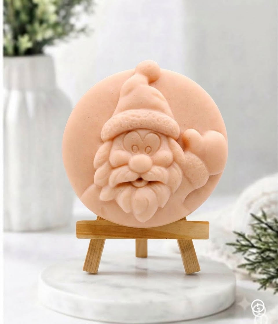 A photograph of a circular, hand-carved soap disc with a relief of Santa Claus’s face, rendered in a pink colour. The soap is presented on a small, light-wood tripod easel, which is positioned on a round white marble pedestal. The background consists of a soft-focus white vase and a white knitted blanket with fir greenery, creating a warm, cosy atmosphere.