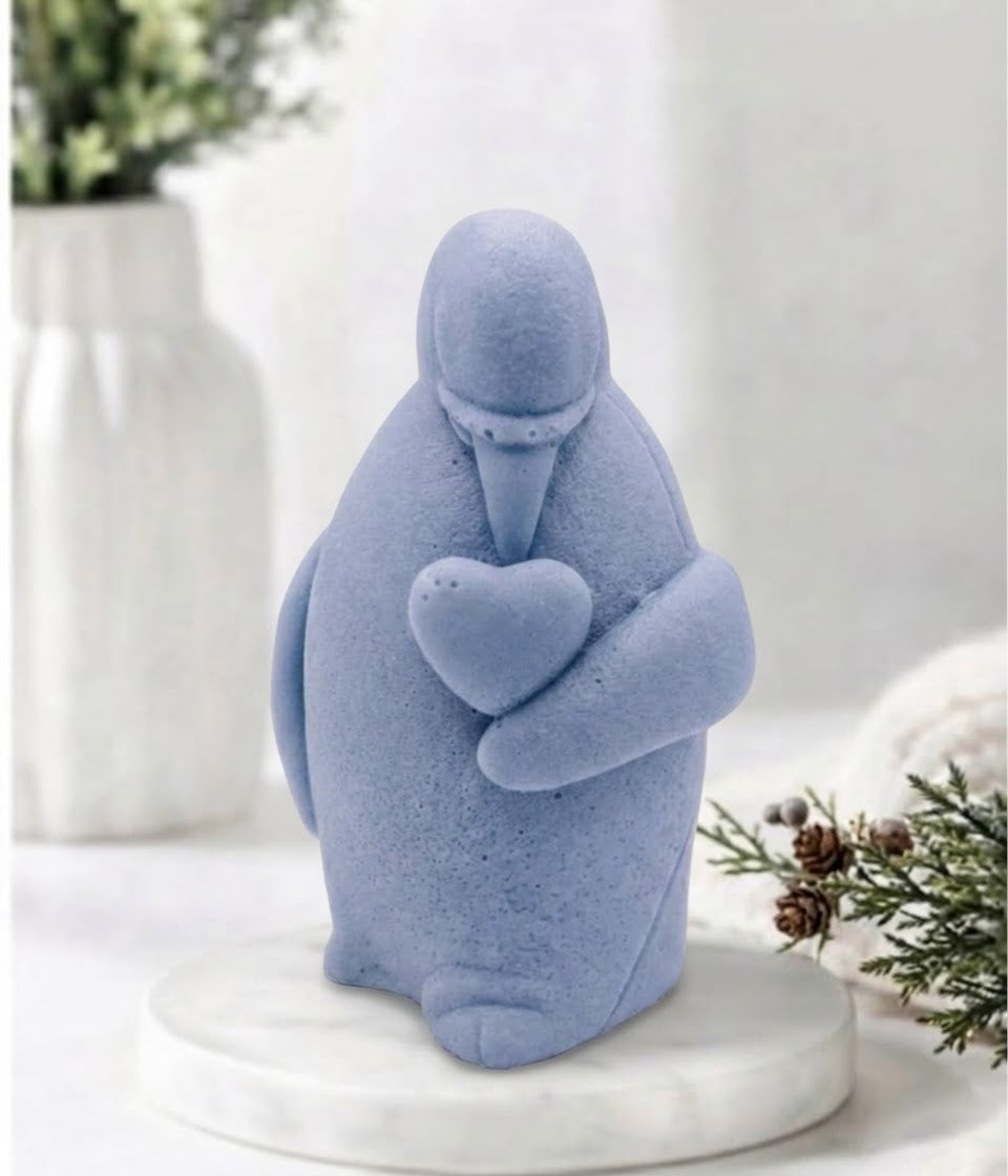 Detailed blue textured penguin artisan soap holding a stone-textured heart, presented on a round marble base with a soft-focus background featuring a white vase and greenery.