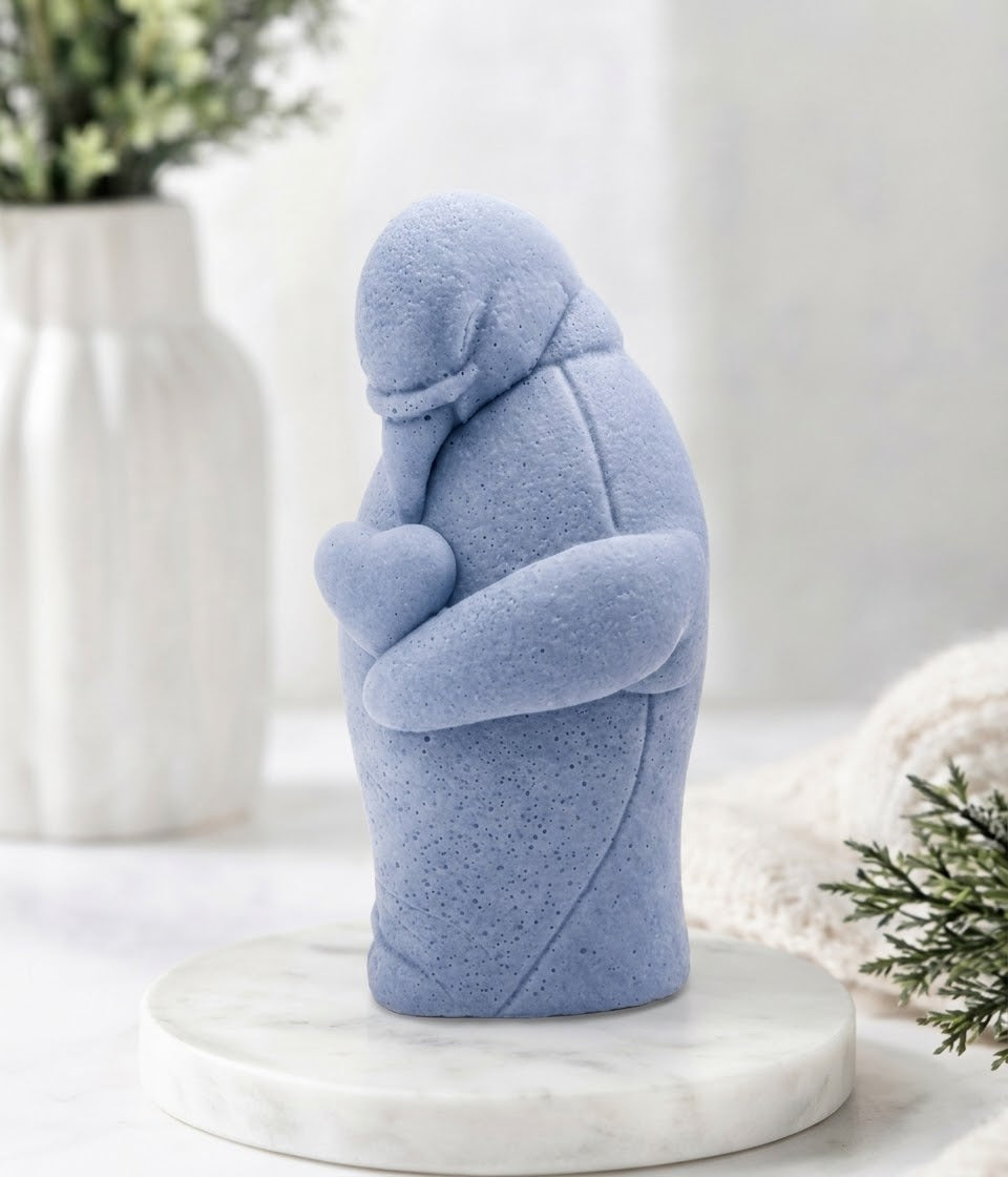 Detailed side view of a blue textured penguin artisan soap holding a stone-textured heart, presented on a round marble base with a soft-focus background featuring a white vase and greenery.