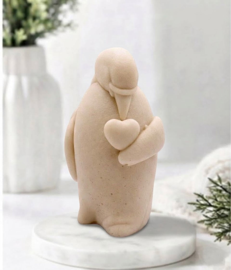 Handmade yellow penguin-shaped artisan soap bar holding a small heart, displayed on a round marble pedestal with a blurred white fluted vase and greenery in the background.