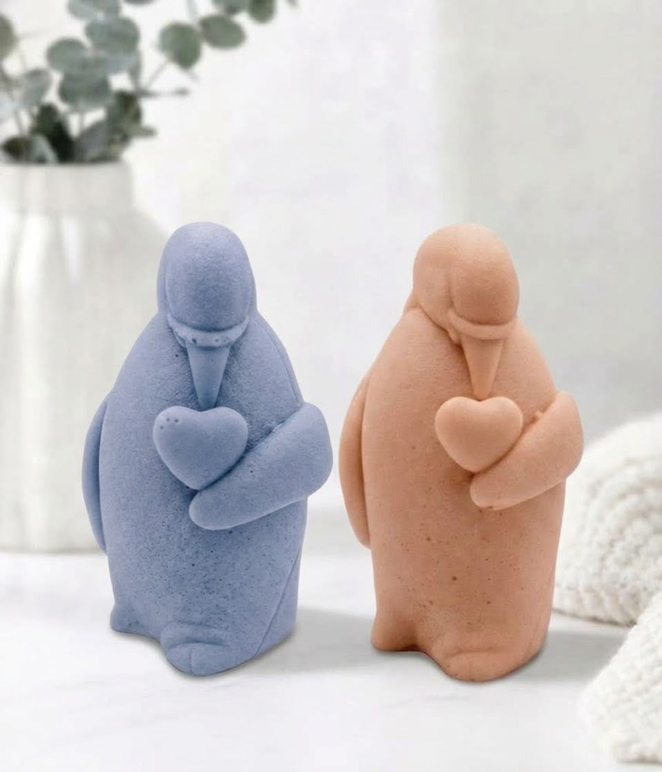 Two artisan penguin-shaped soap bars, one textured blue and one terracotta, stand side-by-side. Both are facing forward, holding their matching stone-textured hearts. The blue penguin holds its heart slightly higher.