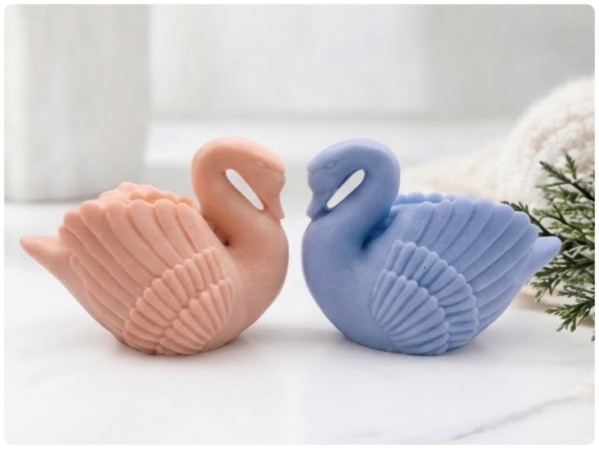 A pair of handmade swan soap bars in soft pink and blue resting together on a textured slab dish.
