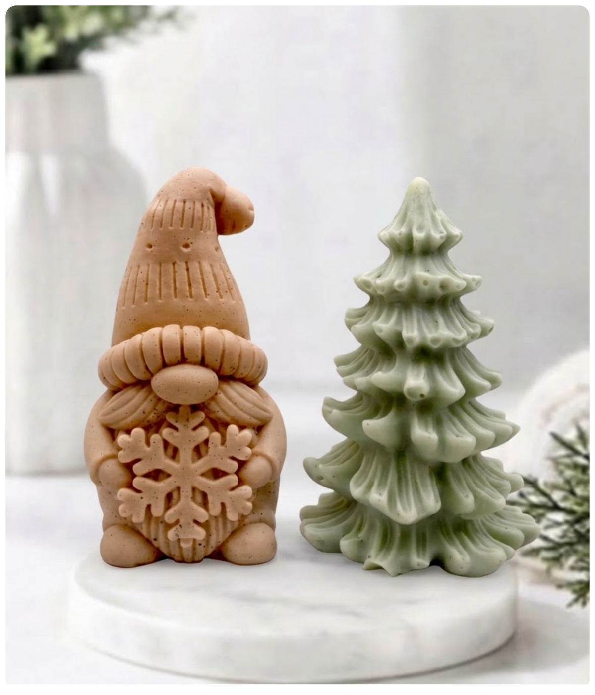 Red clay gnome soap holding a snowflake next to a green Christmas tree soap bar displayed on a marble tray.