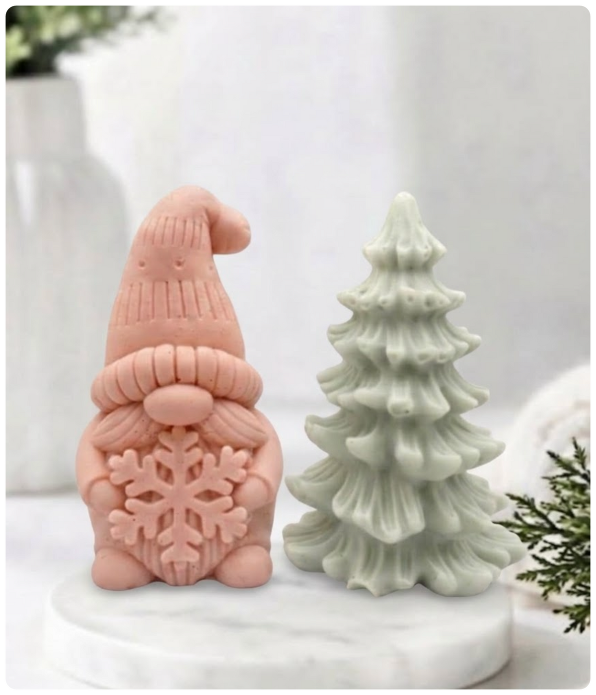Soft pink clay gnome soap holding a snowflake next to a green Christmas tree soap bar on a marble plate.