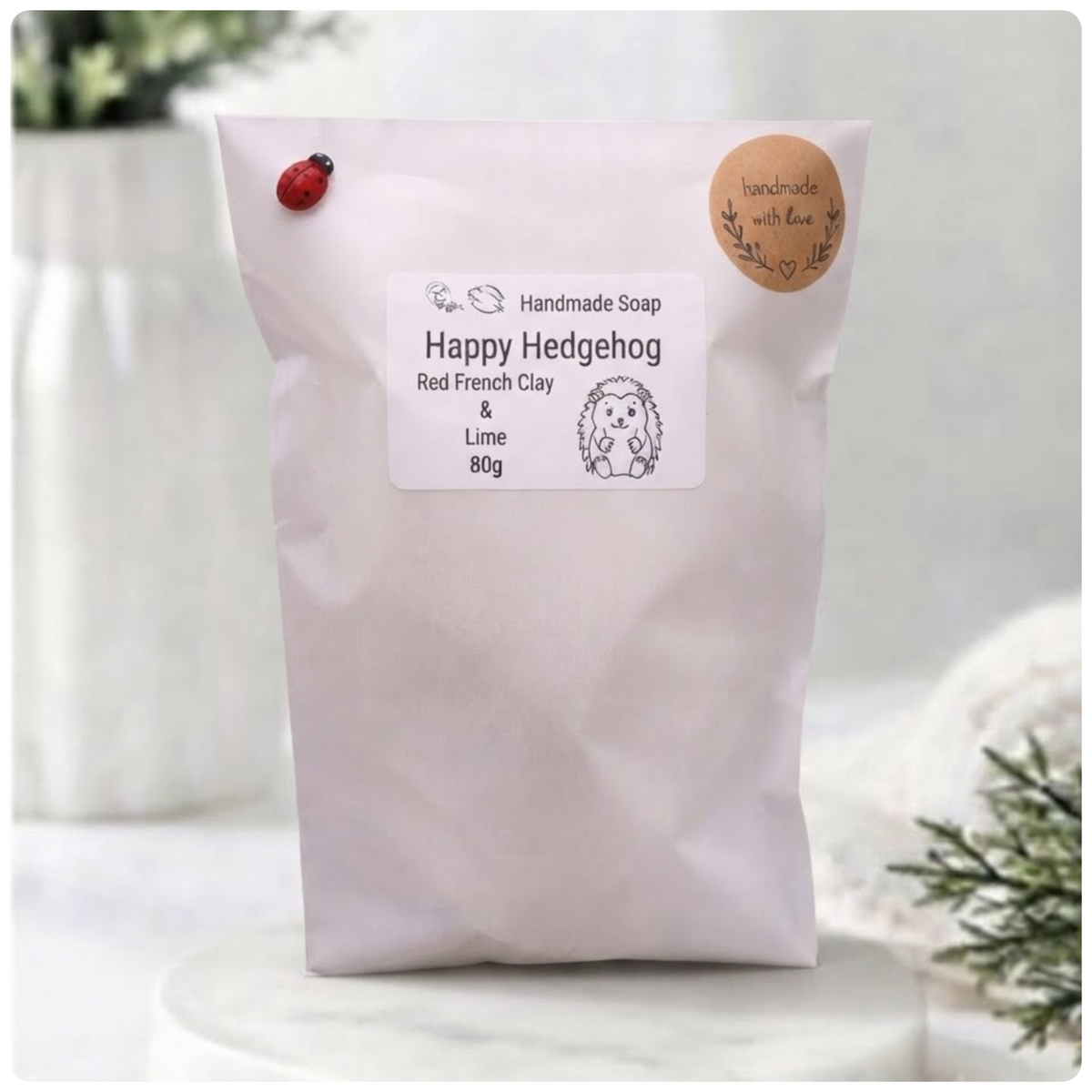A white paper pouch containing 'Happy Hedgehog' handmade soap is placed upright on a white surface. A label on the front describes the soap as 'Happy Hedgehog, Red French Clay & Lime, 80g' and features a cartoon hedgehog. A small ladybug is pinned to the top left of the pouch, and a round 'handmade with love' sticker is on the top right. The background is a blurred bathroom scene.