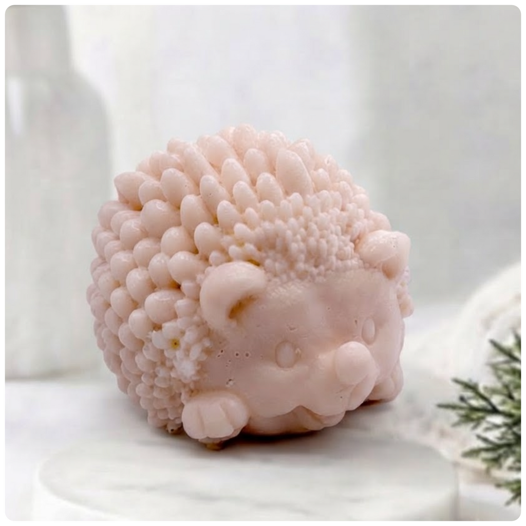A light pink, sculpted hedgehog soap figure, detailed with small spikes and a smiling face, sits on a white marble surface against a blurred bathroom background.