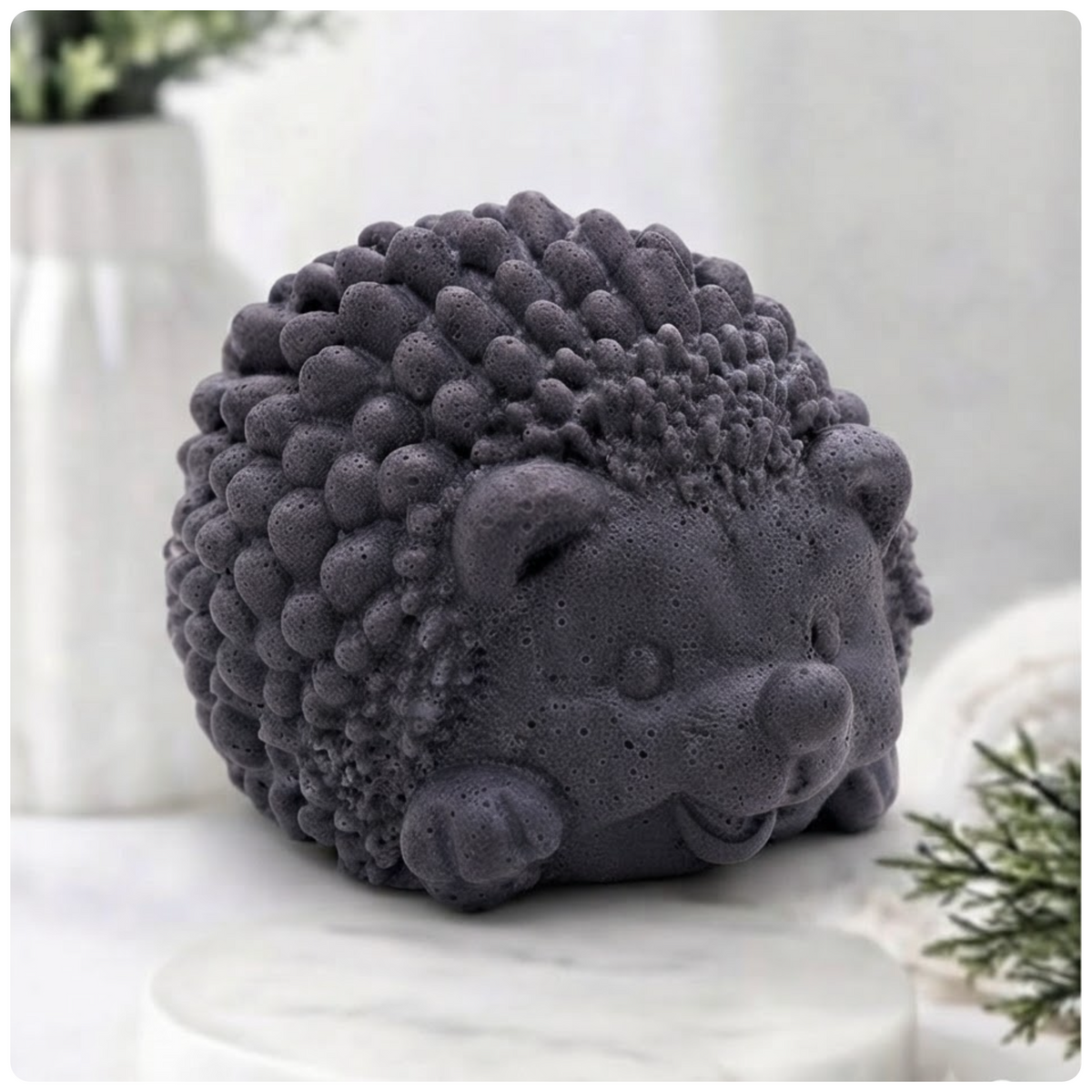 A dark grey, textured hedgehog soap figure, detailed with small spikes and a smiling face, sits on a white marble surface against a blurred bathroom background.