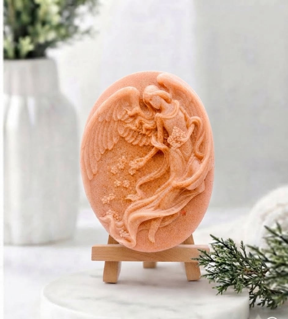 An oval terracotta coloured soap bar featuring a detailed relief carving of an angel, displayed on a small wooden stand on a white marble surface with a blurred background.