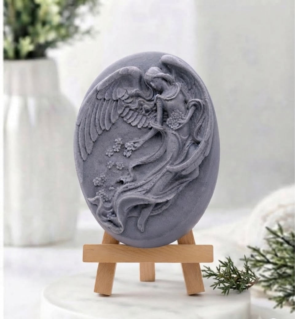 A gray charcoal soap bar featuring a detailed relief carving of an angel with wings and flowing robes, displayed on a small wooden easel stand over a white marble surface.