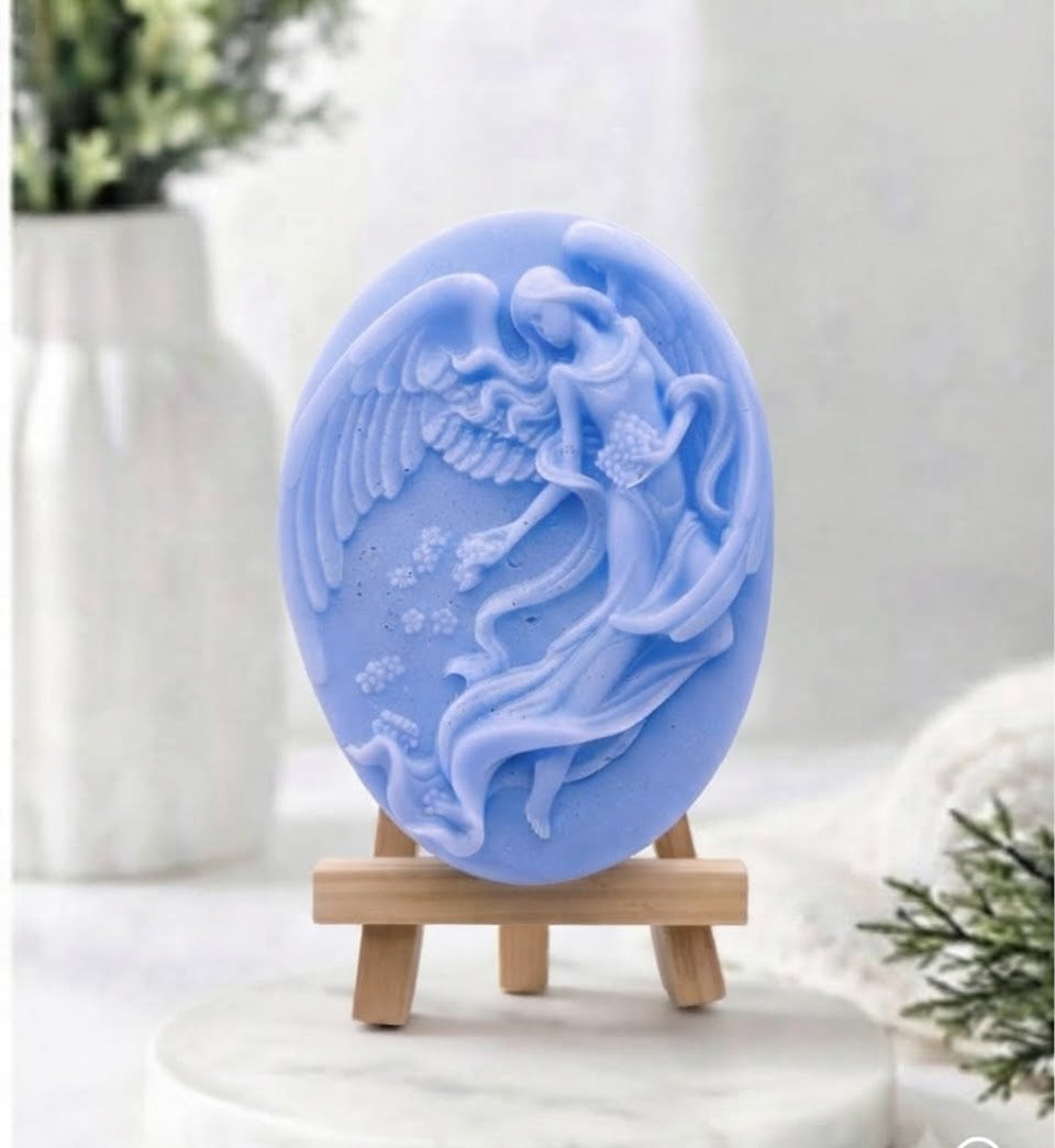 A light blue oval soap bar with a raised winged figure design, displayed on a small wooden stand on a white marble surface with a soft blurred background 