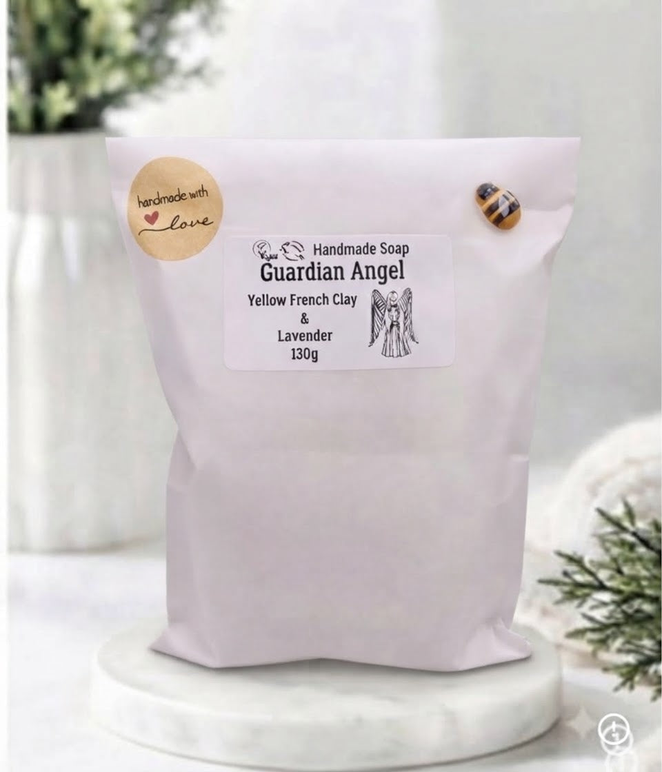 A white paper pouch containing handmade soap stands on a wooden surface against a blurred bathroom background. The pouch has a label that reads "Handmade Soap Guardian Angel, Yellow French Clay & Lavender, 130g," along with an angel illustration. A round sticker to the left reads "Handmade with Love," and a small bee charm is pinned to the top right.