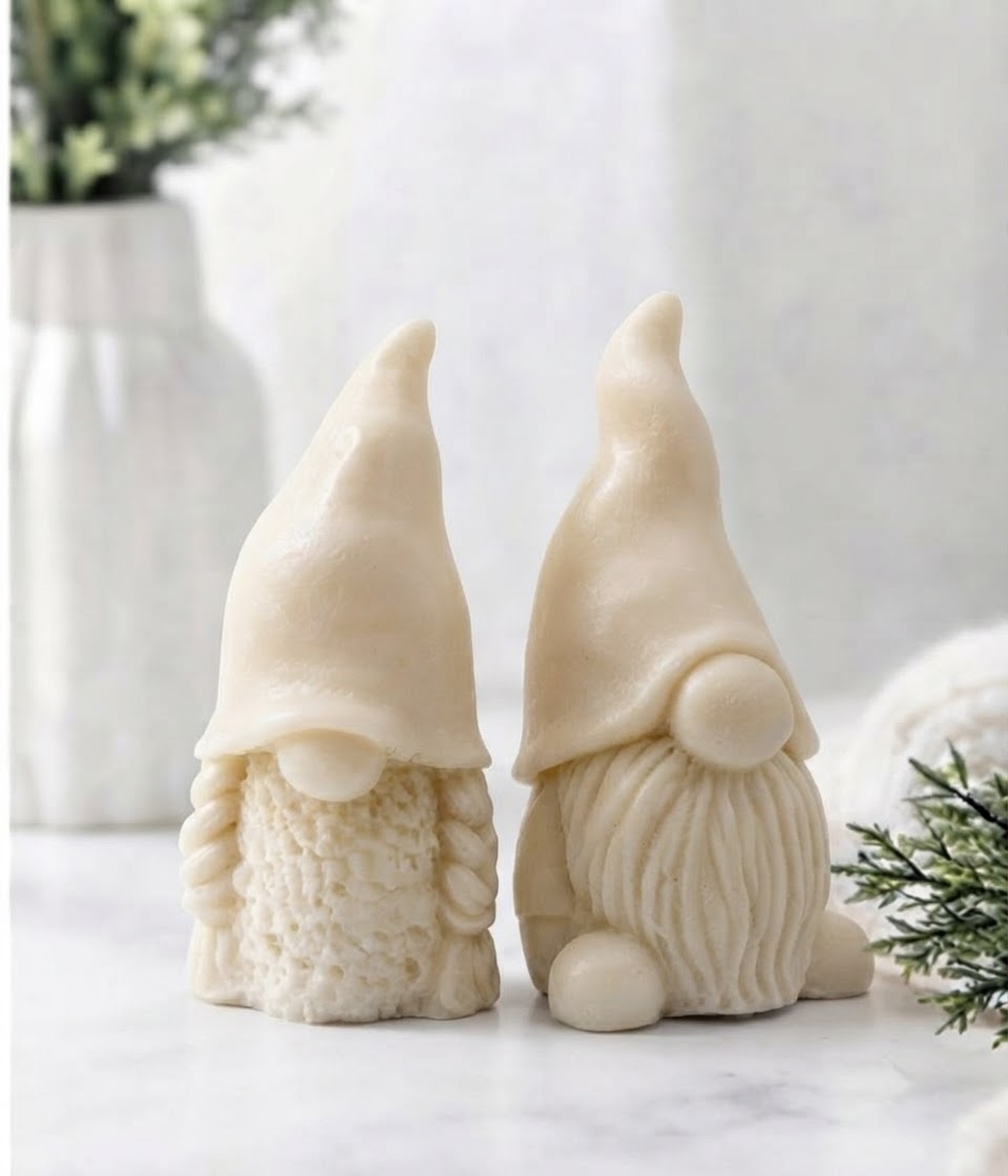 Two gnome soaps in a yellow tone, standing side by side on a light marble-like surface with soft blurred decorative plants in the background.
