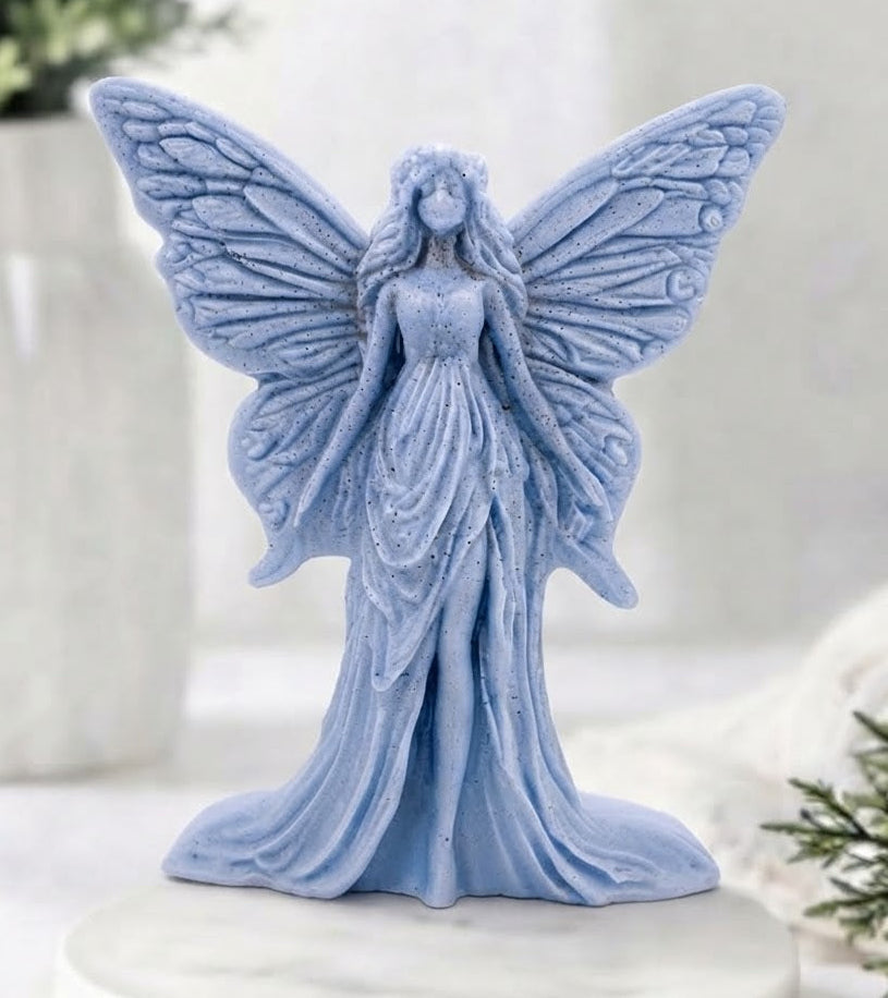 A light blue winged fairy vegan soap with a long flowing gown, standing on a round white marble-style base against a softly blurred indoor background with plants.