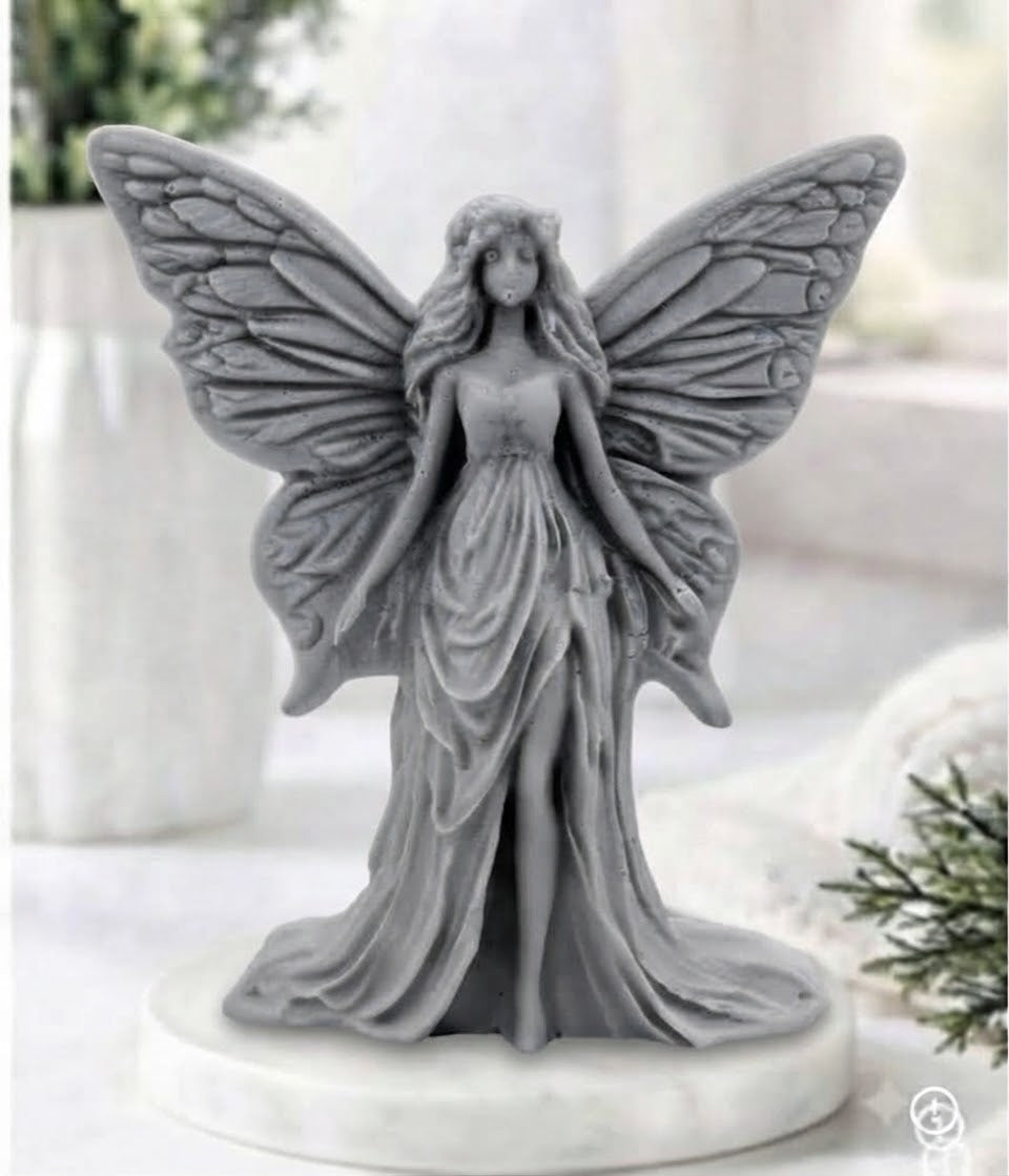 A charcoal gray winged fairy vegan soap with detailed wings and a flowing gown, standing on a round white base in a softly blurred indoor setting.
