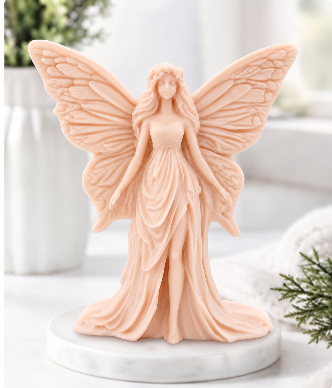 A soft pink coloured angel figurine with large wings and a flowing gown, standing on a round white marble base in a softly blurred indoor setting.