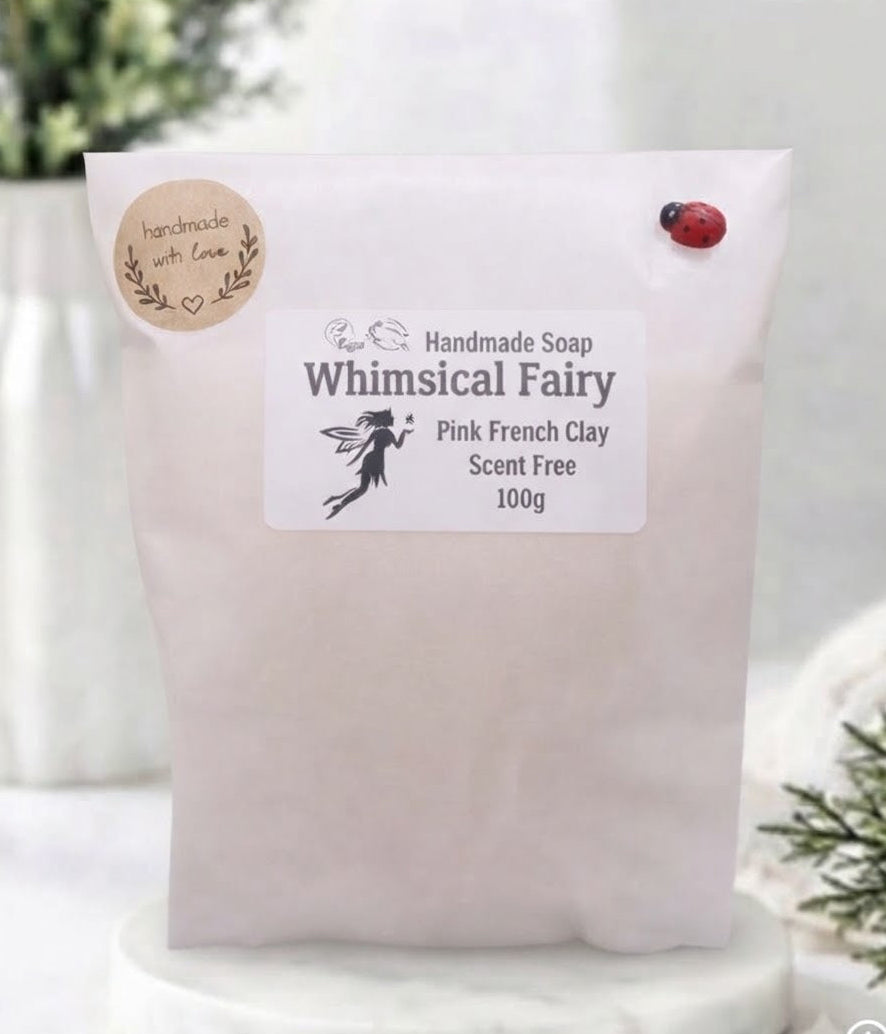 A 100g pouch of Whimsical Fairy Pink French Clay handmade soap, displayed on a white marble coaster with a soft, blurred background of greenery and a vase.
