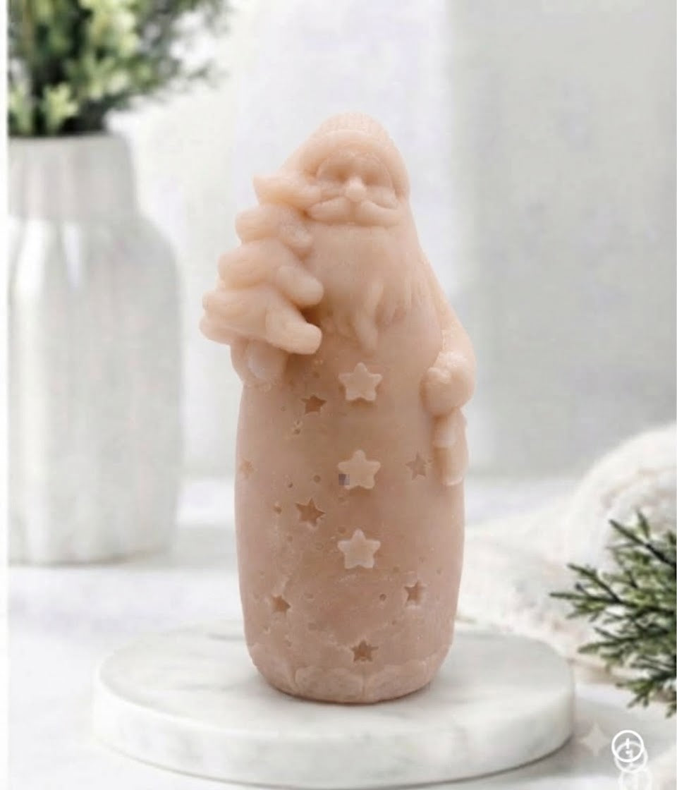 A pale blush pink Santa Claus vegan soap with star cutouts, displayed on a white marble coaster with a soft-focus background of a white vase and greenery.