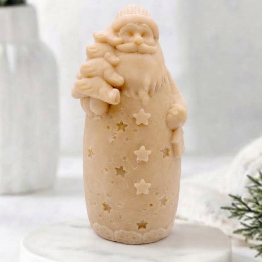 A yellow Santa Claus vegan soap holding a Christmas tree, standing on a white marble round plinth against a soft, bright, blurred background with a white vase and greenery.