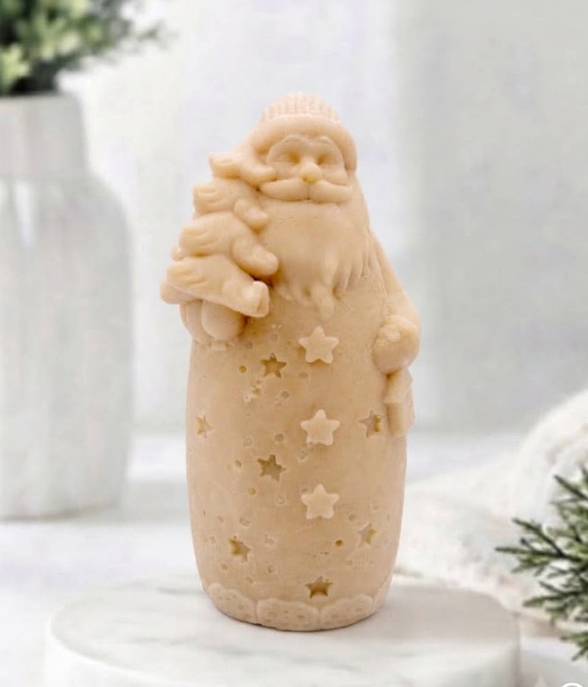 A yellow Santa Claus vegan soap holding a Christmas tree, standing on a white marble round plinth against a soft, bright, blurred background with a white vase and greenery.