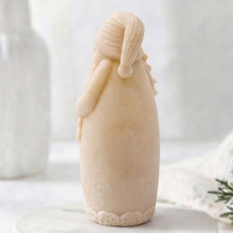 A yellow coloured rear view of Santa Claus vegan soap standing on a round white marble plinth, with a soft-focus background featuring a white vase with greenery and a white knit blanket.