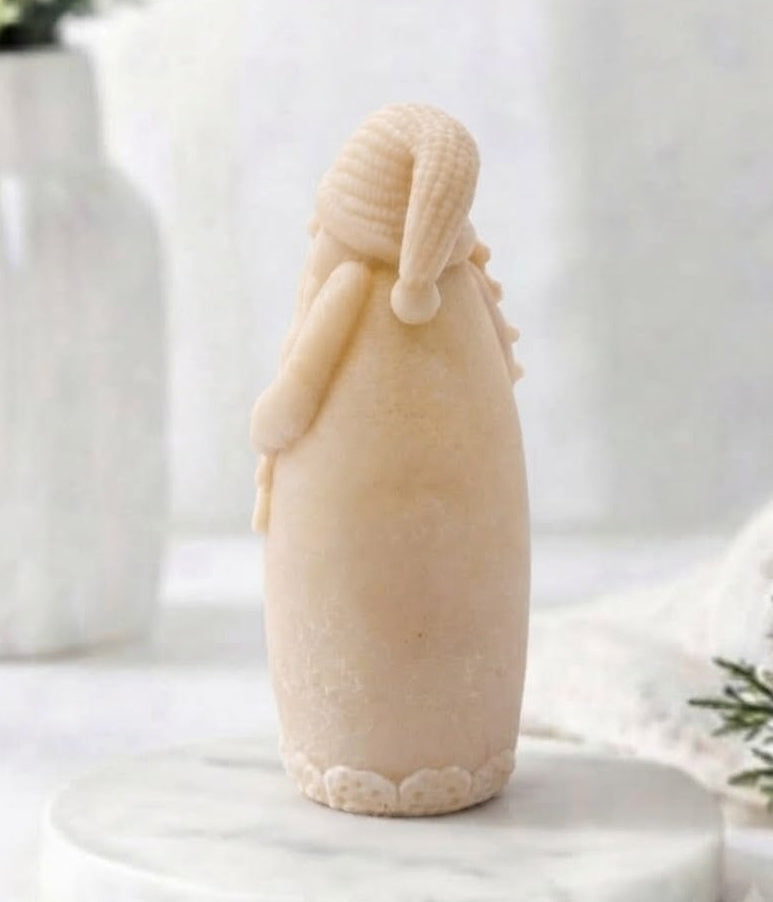 A yellow coloured rear view of Santa Claus vegan soap standing on a round white marble plinth, with a soft-focus background featuring a white vase with greenery and a white knit blanket.