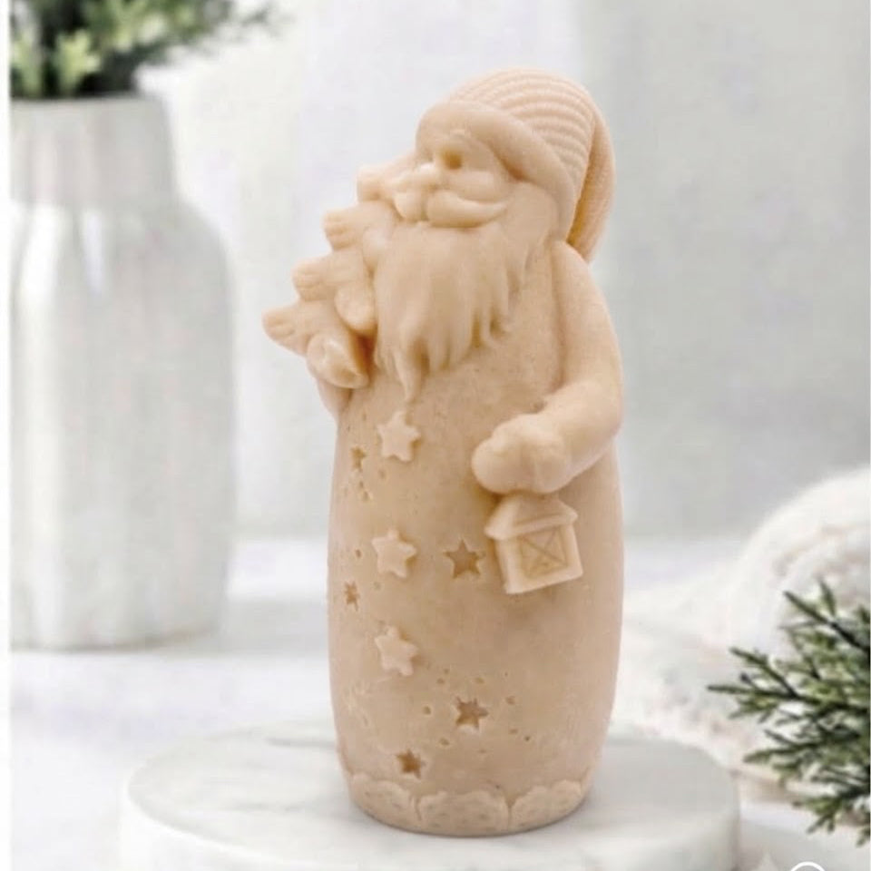 A yellow Santa Claus vegan soap standing on a round white marble base, holding a sack and a small lantern, photographed in a softly styled indoor setting with blurred greenery and white decor in the background.
