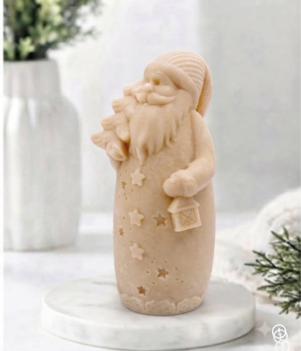 A yellow Santa Claus vegan soap standing on a round white marble base, holding a sack and a small lantern, photographed in a softly styled indoor setting with blurred greenery and white decor in the background.