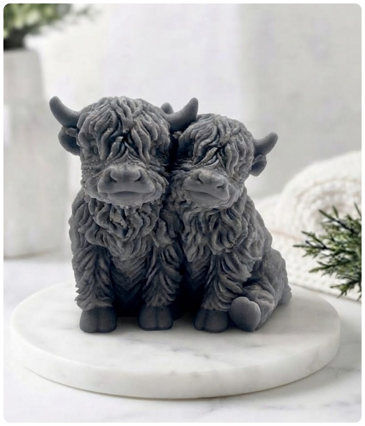 Two charcoal  grey highland cow vegan soap with shaggy fur sitting side by side on a round white marble platter, set against a softly blurred neutral background with small green plant accents.