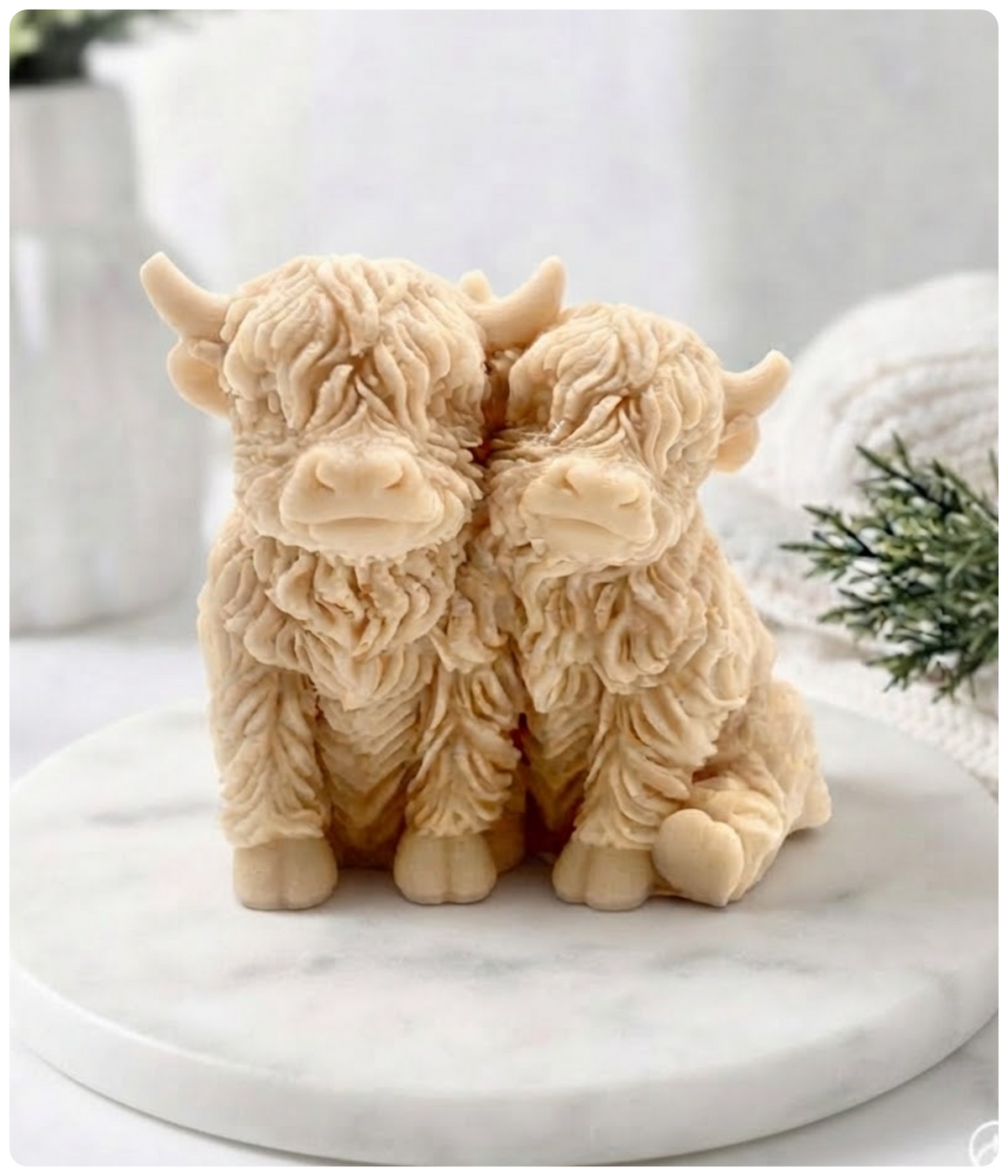 Two yellow Highland cow vegan soaps with textured fur, sitting side by side on a round white marble platter against a softly blurred background.