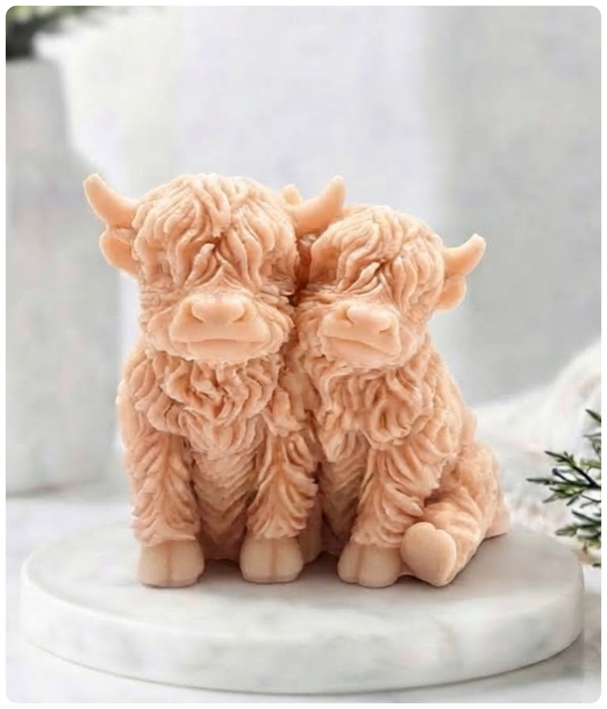 Two pink nuzzling Highland cow soap in a warm peach-beige tone, posed together on a round white marble base against a soft light gray background.