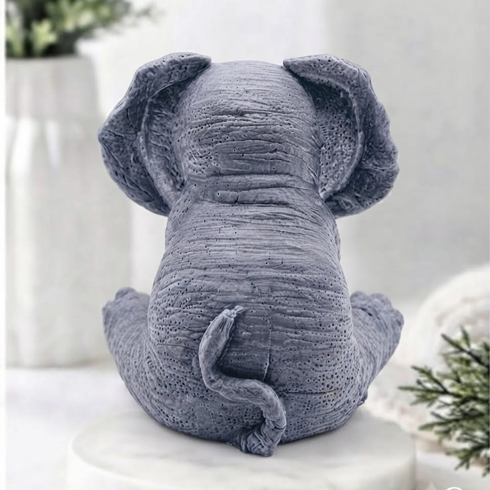 A gray elephant vegan soap sitting on a round white marble base, photographed in a softly styled indoor setting with blurred plants in the background.