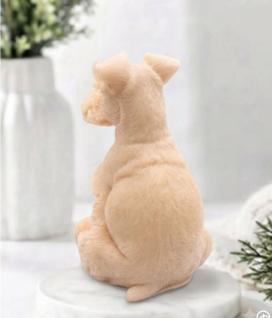 A rear view of a soft pink schnauzer dog soap sitting on a round white marble base, photographed in a softly styled indoor setting with blurred plants and neutral decor