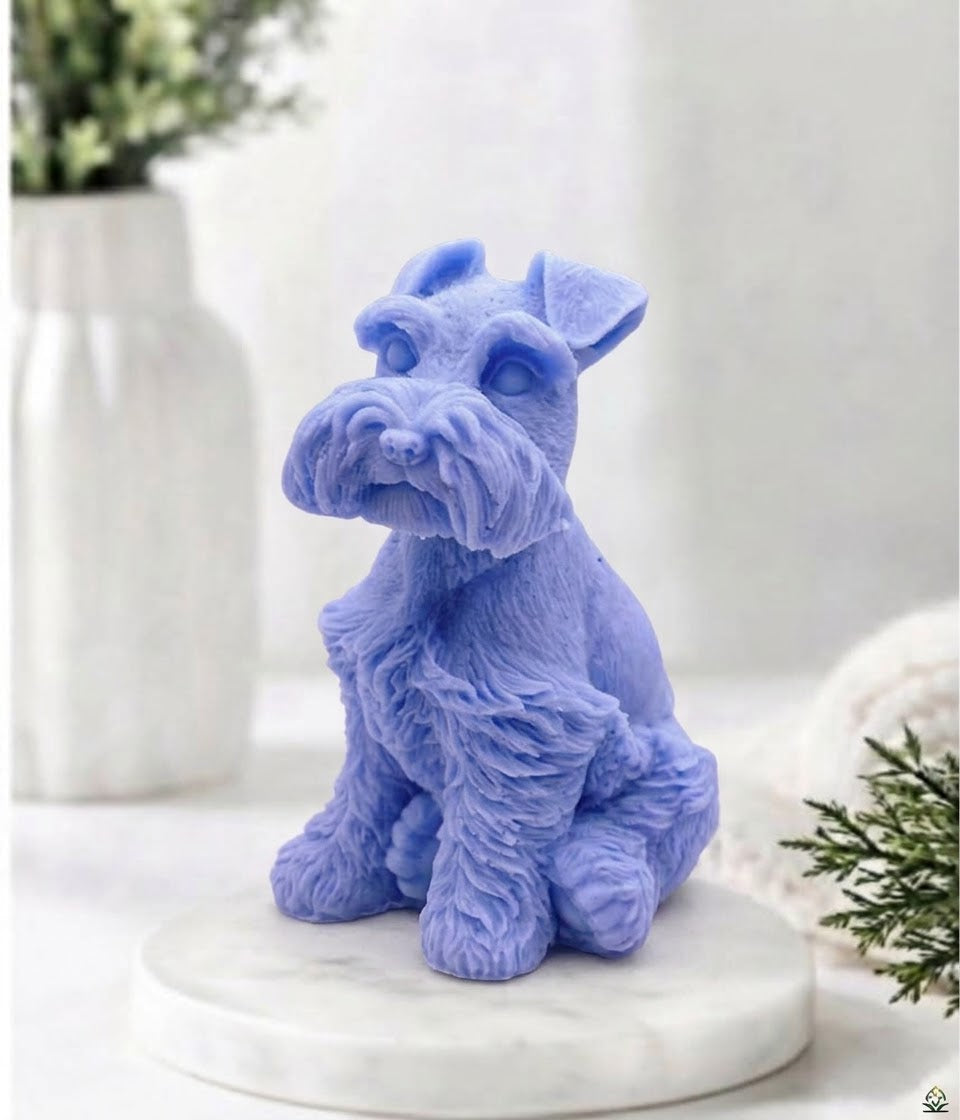 A lavender-blue vegan dog soap, resembling a Schnauzer, sitting on a round white marble base. The background is a softly blurred, bright white interior with a white vase and green foliage.