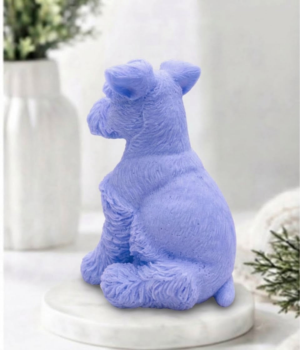 A lavender blue dog-shaped soap gift with textured fur, sitting on a white marble coaster with a blurred white vase and greenery in the background.