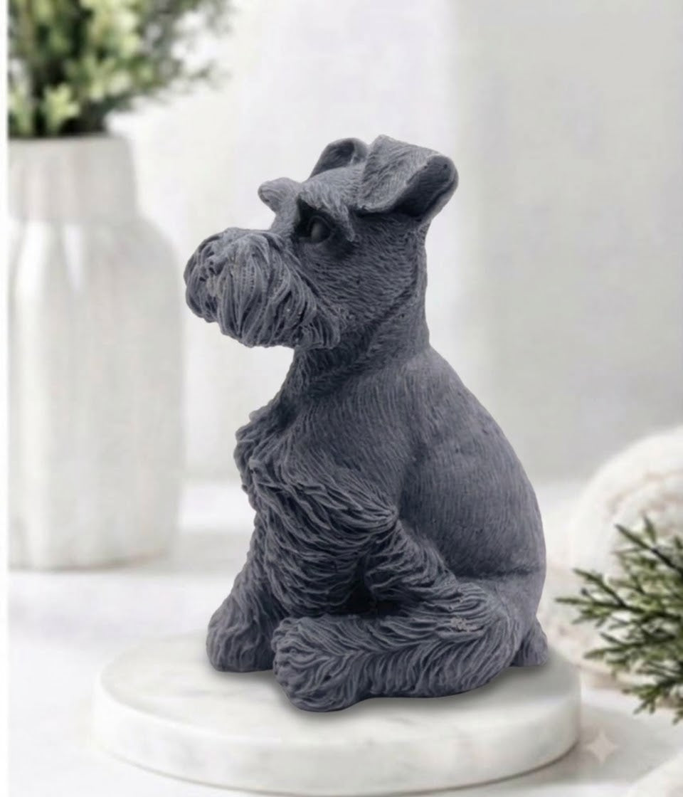 A dark charcoal gray vegan soap of a Schnauzer dog sitting on a round white marble base, styled on a white surface with soft-focus decorative plants in the background.