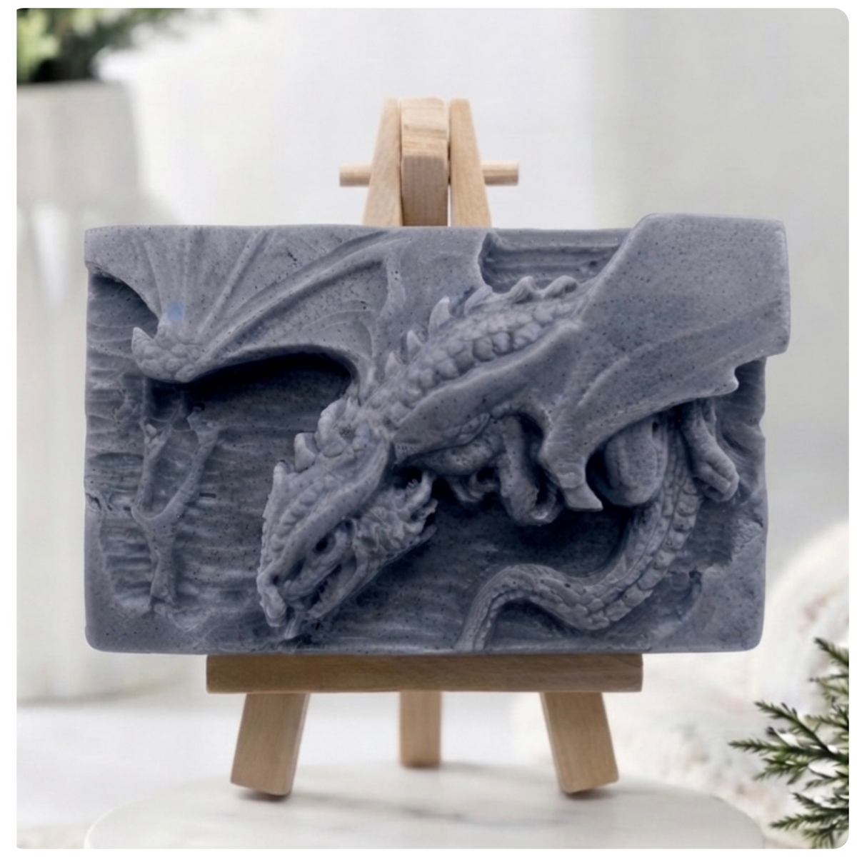 A gray rectangular relief vegan soap featuring a detailed dragon carving, displayed on a small wooden easel against a softly blurred indoor background.