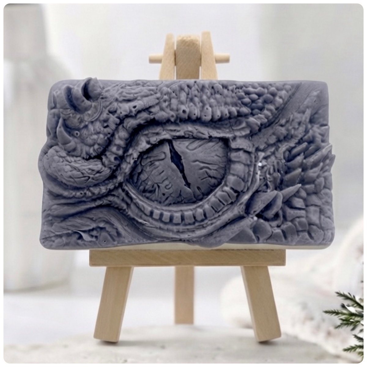 A grey rectangular vegan soap featuring a detailed dragon eye design, displayed on a small wooden easel against a soft, blurred white background.
