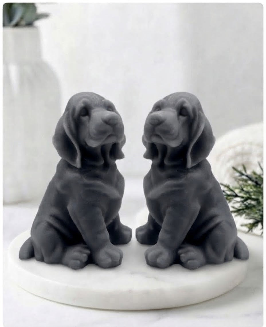 Two charcoal grey bloodhound soaps sitting on a white marble display stand with a soft bathroom background.