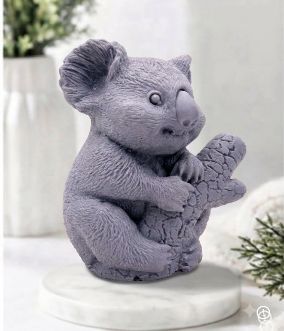 A side-facing photograph of a grey, charcoal-coloured koala-shaped soap bar. The highly detailed soap is sculpted to show textured fur and the koala clinging to a textured log. It sits on a round, white marble disc. In the blurry background, there is a white ceramic vase with green sprigs and a rolled white towel.