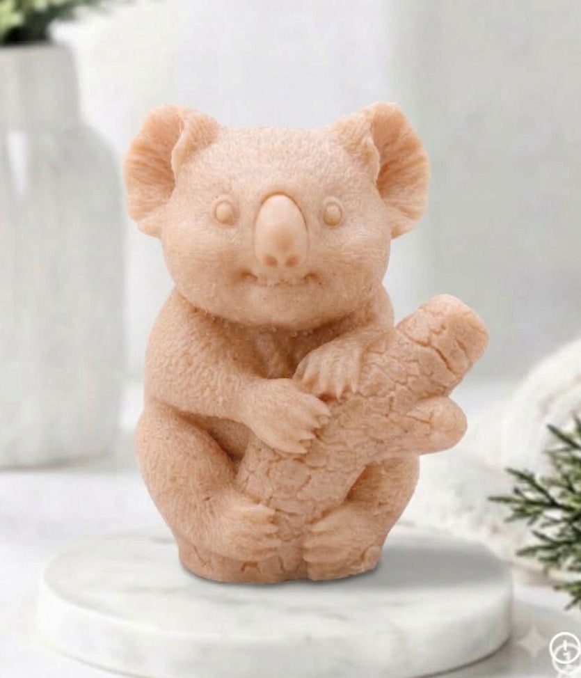 A front-facing photograph of a koala-shaped soap bar in a soft-pink French clay. The detailed soap is sculpted with a soft fur texture and the koala holding a small log. It sits on a round, white marble disc. The blurred background shows a white vase with eucalyptus sprigs and a rolled white towel.