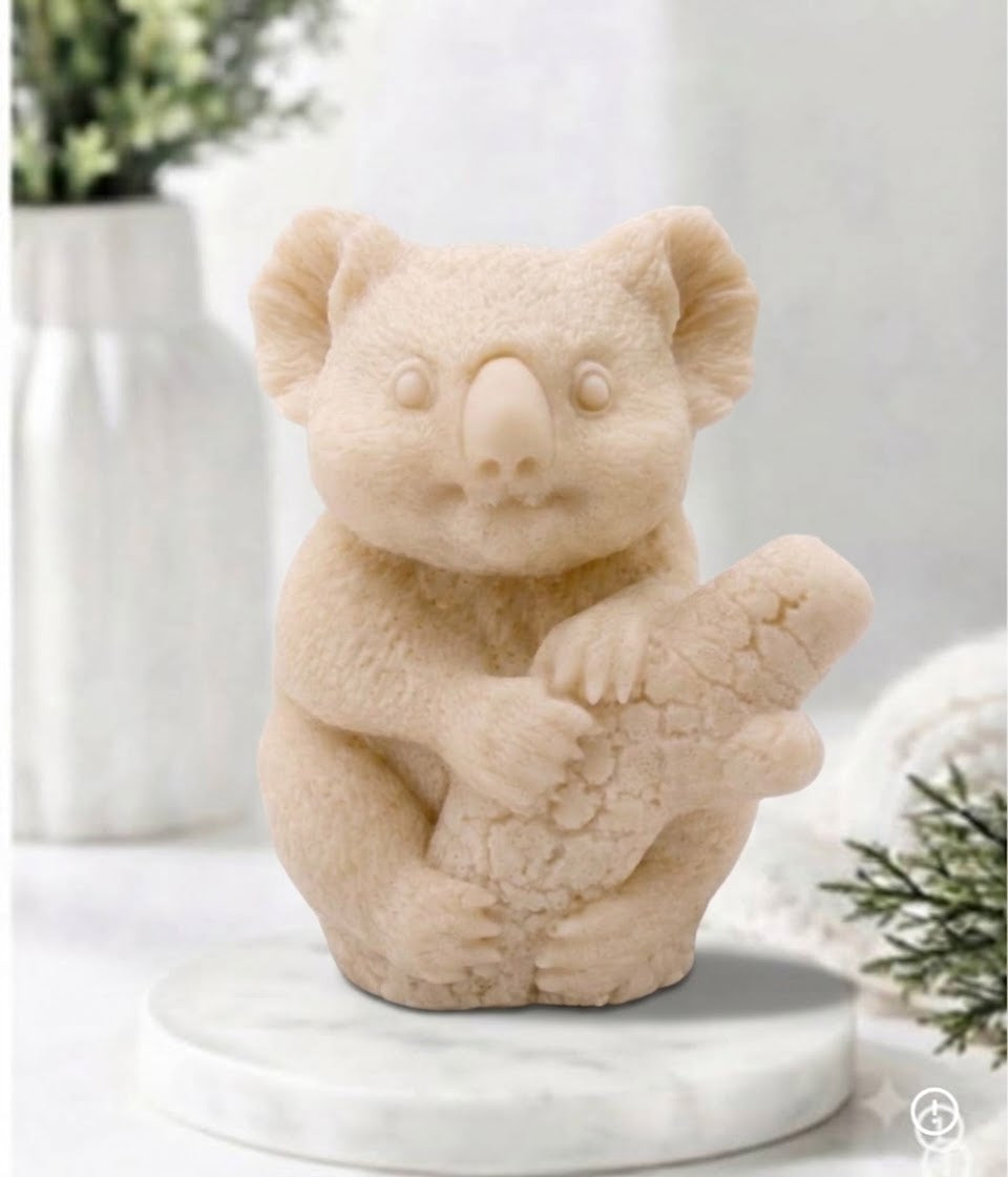 A front-facing photograph of a yellow French clay, koala-shaped soap bar. The detailed sculpture shows fur texture and the koala clinging to a log. It sits on a round, white marble disc. The blurred background features a white vase with green sprigs and a rolled white towel.