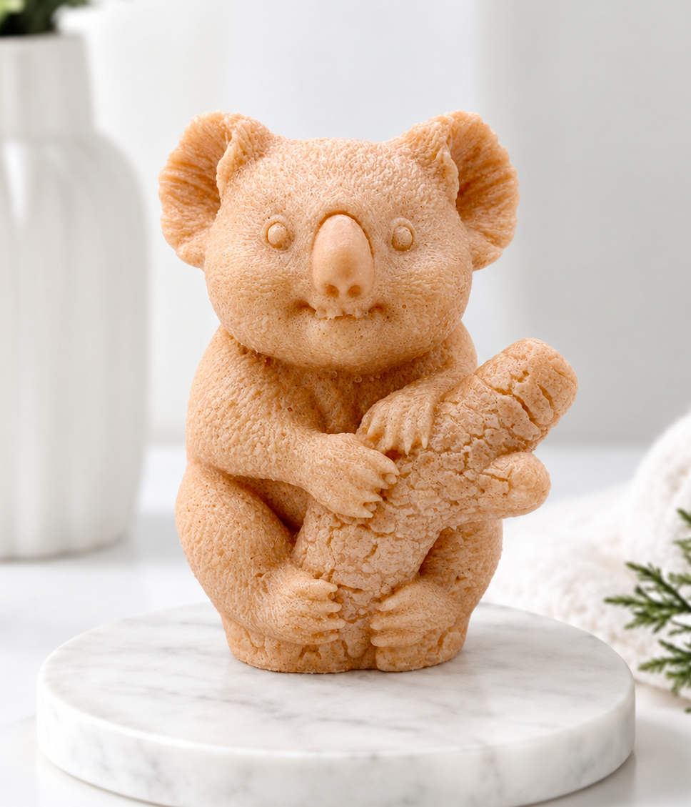 A front-facing photograph of a koala-shaped soap bar in a red French clay terracotta shade. The detailed soap is sculpted with a fuzzy fur texture and the koala holding a log. It sits on a round, white marble disc. In the blurry background, there is a white vase with greenery and a rolled white towel.